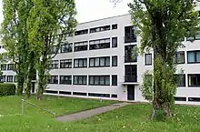 Photographie couleur d’une façade d’immeuble d’habitation. Au premier plan, verdure et arbres.