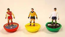 Photo serrée de trois mini-figurines représentant des footballeurs du jeu Subbuteo, une rouge, une jaune et un vert.