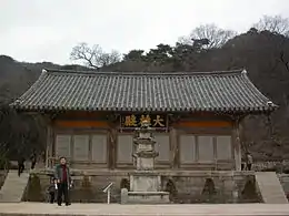Photographie de l'extérieur du bâtiment d'un temple. Il est derrière une statue, et trois grands caractères chinois surplombent une porte.