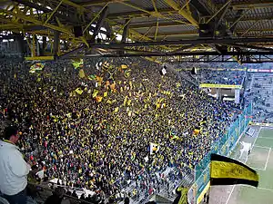 Vue depuis sous les toits d'une tribune à gauche pleine de supporters debout avec des drapeaux jaunes en plein match.