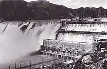 Photographie d'un barrage vu de loin. Des trombes d'eau s'échappent par le sommet du barrage. Au premier plan un grand bâtiment de plusieurs étages est accolé à ce barrage.