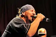 Photo de Mike Muir, chanteur de Suicidal Tendencies, le groupe fait partie de la bande son. Sur cette photo prise du côté droit, il porte un T-shirt sombre et un bandana sur le front et un micro dans la main droite, alors qu'il est en train de chanter.