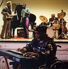 Sun Ra au premier plan das un fauteuil roulant, en train de jouer sur un clavier. Derrière, une scène avec des musiciens.