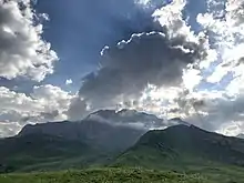 Un splendide nuage couronne une haute montagne recourverte d'herbe verte.