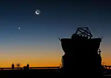 Coucher de soleil derrière un grand télescope. Des personnes regardent l'horizon, où l'on voit un point lumineux et la Lune.