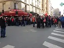 Photo montrant des personnes rassemblées dans la rue devant une enseigne rouge et noire.