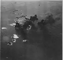 Photographie aérienne noir et blanc quasi verticale d'une île d'où s'élèvent des nuages de fumée noire. Un avion est visible au milieu des fumées.