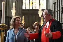 Photo d'un ecclésiastique en soutane rouge recevant une dame blonde en tailleur bleu.