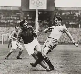 Photographie en noir et blanc d'une scène d'un match de football : un joueur en blanc tacle l'autre, en noir.