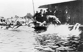 Plusieurs hommes en maillot en train de plonger dans l'eau depuis un ponton où se trouvent d'autres hommes en costume.