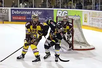 Deux hockeyeurs au premier plan, penchés, la tête tournée vers leur gauche. Au second plan, un gardien de but.