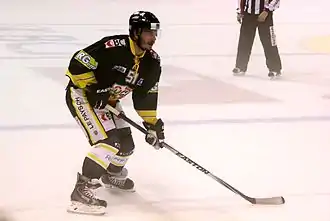 Hockeyeur, de trois quarts face, légèrement penché en avant, la canne entre ses deux mains et posée sur la glace