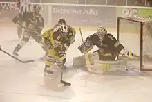 Dans la brume, un hockeyeur penché en avant au premier plan, un gardien de but sur sa gauche, un joueur caché et à l’arrière-plan, un quatrième joueur et un arbitre