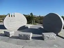 Photo en plan large des deux pierres commémoratives, présentant des inscriptions en français et en anglais, avec trois petits bancs de pierre en face d'elles, le tout formant un arc de cercle.
