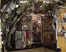 Photo couleur prise à l'intérieur d'une reconstitution de l'avion dans un hangar. Les débris métalliques sont placés sur des grilles correspondant à leur emplacement sur l'avion.