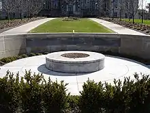 Photo d'un monument circulaire en pierre comportant une plus petite partie ronde au centre ainsi qu'un mur, situé quelques mètres plus loin, l'entourant. Á l'arrière-plan se trouve de l'herbe, entouré des deux côtés par un chemin en pierre.
