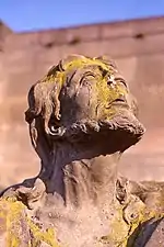 Détail d'une sculpture, tête d'homme barbu levée vers le ciel et envahie par la mousse.