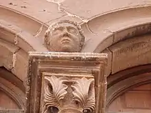 Photo couleur présentant le chapiteau d'une colonne. Décoré de feuillages, il est surmonté d'une tête à la jointure des arcs brisés qui en partent.