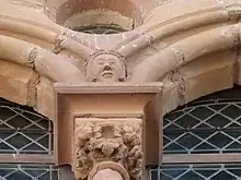 Photo couleur montrant un chapiteau de fenêtre sculpté de feuilles de vigne et surmonté d'une tête humaine et d'un oculus. Les ouvertures sont dotées de panneaux de vitraux de verre en losange.