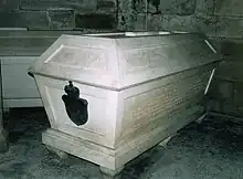 Photo en couleurs d'un sarcophage en marbre blanc portant un blason en bronze sur la face principale.