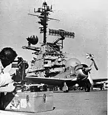 Photographie en noir et blanc d’un jeune homme au premier plan en train de filmer un porte-avions.