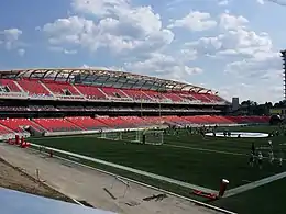 Un terrain de sport vert, des gradins rouges et un ciel bleu comprenant quelques nuages.