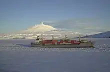 Photographie d'un bateau traversant la banquise, et en arrière un volcan fumant, couvert de neige.