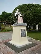 Statue femme âgée assise. Longue jupe rose clair, haut clair à manches longues, visage émacié, cheveux tirés en arrière.