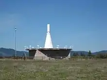 Antenne blanche en forme de cône allongé surmontant un bâtiment métallique entouré d'une barrière en bois.