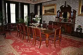 La table de salon et ses douze chaise est richement ornée. L'intérieur de la pièce comporte une décoration abondante faite de meubles et de tableaux. Un grand tapis sur lequel se trouve la table orne le sol