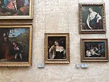 Photo couleur d’un mur de couleur crème sur lequel sont accrochés cinq tableaux (deux à gauche (l’un figure un homme assis et un jeune agenouillé devant lui ; seule la partie en bas à droite de l’autre est visible), un au centre (portrait d’une femme) et deux à droite (portrait d’une femme et peinture de deux chiens)).
