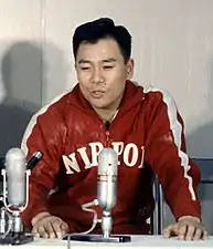 Un homme avec une veste en cuir rouge sur laquelle est écrit Nippon parlant dans un micro.