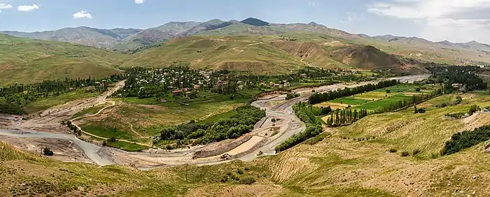 Devant une chaîne de montagnes basses au lointain, auprès d'une courbe dans une rivière, un bourg au milieu de champs d'agriculture extensive.