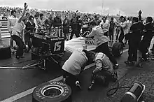 Photographie en noir et blanc d'une monolace de Formule 1, vue de derrière, dans la voie des stands, entourée de personnes.