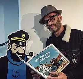 Photographie d'un homme portant des lunettes et un chapeau, lisant une bande dessinée qu'il tient dans les mains, devant un mannequin en carton à l'effigie du capitaine.