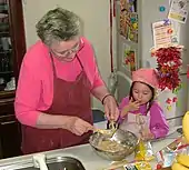 Une grand-mère portant un tablier bordeaux sur une blouse rose, racle au-dessus du plat en inox de pâte qu'elle vient de confectionner l'un des fouets de son mixeur-batteur tandis qu'à côté d'elle sa petite-fille habillée de rose fuchsia et portant un tablier identique mais rose pâle, se lèche le doigt utilisé pour gouter la pâte prélevée sur le second fouet.