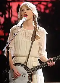 Taylor Swift, wearing a strapless dress, looks directly at the camera. Her hair is tied back, with a few curly tendrils loose