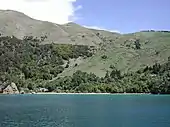 Photographie d'un littoral très pentu et déboisé en partie haute, au pied duquel quelques maisons et un ponton sont visibles.