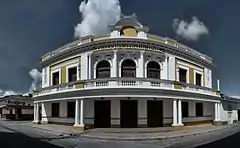 Photo en couleurs d'un bâtiment à deux étages, de couleur blanche et jaune ocre, avec des colonnes et ornements néoclassiques