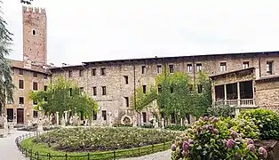 Une cour remplie de jardins ; sur les bords des statues ; au fond des bâtiments en pierres, certains sont couverts de lierre ; au fond à gauche une tour crénelée.