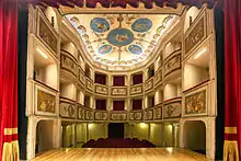 L'intérieur d'une petite salle richement décorée et fortement illuminée, dans les couleurs rouge et or ; un parterre et deux galeries en hauteur ; le plafond est peint.