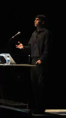Photo d'un homme debout, habillé en noir, face à un micro en train de faire une conférence.