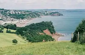 Vue depuis une falaise dominant la baie ; vaste plage, petite agglomération au-dessus du rivage ; ciel bleu avec quelques nuages épars.