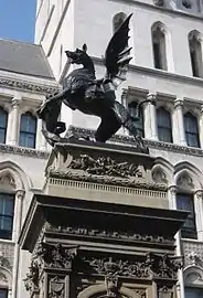 Photo. Griffon ailé en bronze vu de profil, tenant un écu, sur haut socle carré sculpté dont seul le haut est visible ; immeubles cossus en fond