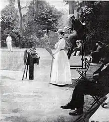 Vue d'un terrain de tennis avec une femme en robe blanche tenant une raquette et un arbitre sur sa chaise.