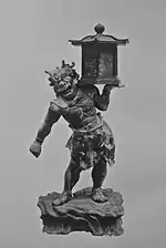  Tentōki. Vue de face d'une statue trapue avec un visage de démon. Elle porte une lanterne sur l'épaule gauche qu'elle soutient de sa main gauche. Photographie noir et blanc.