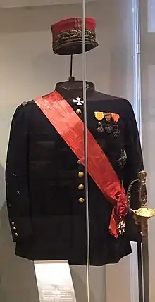 Photographie d'un uniforme noir, une écharpe rouge le traversant. Des décorations sont visibles sur le torse. Il est surmonté d'un képi rouge.