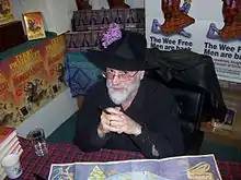 Terry Pratchett à 57 ans, vêtu de noir et avec un chapeau, posant à un stand.