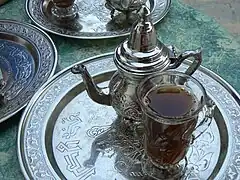 Une théière, un verre rempli de thé à la menthe et un plateau argenté avec des inscriptions en arabe.
