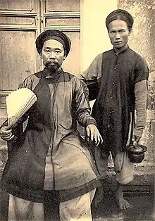 Photographie de deux hommes asiatiques en habit traditionnel, l'un assis et l'autre debout.